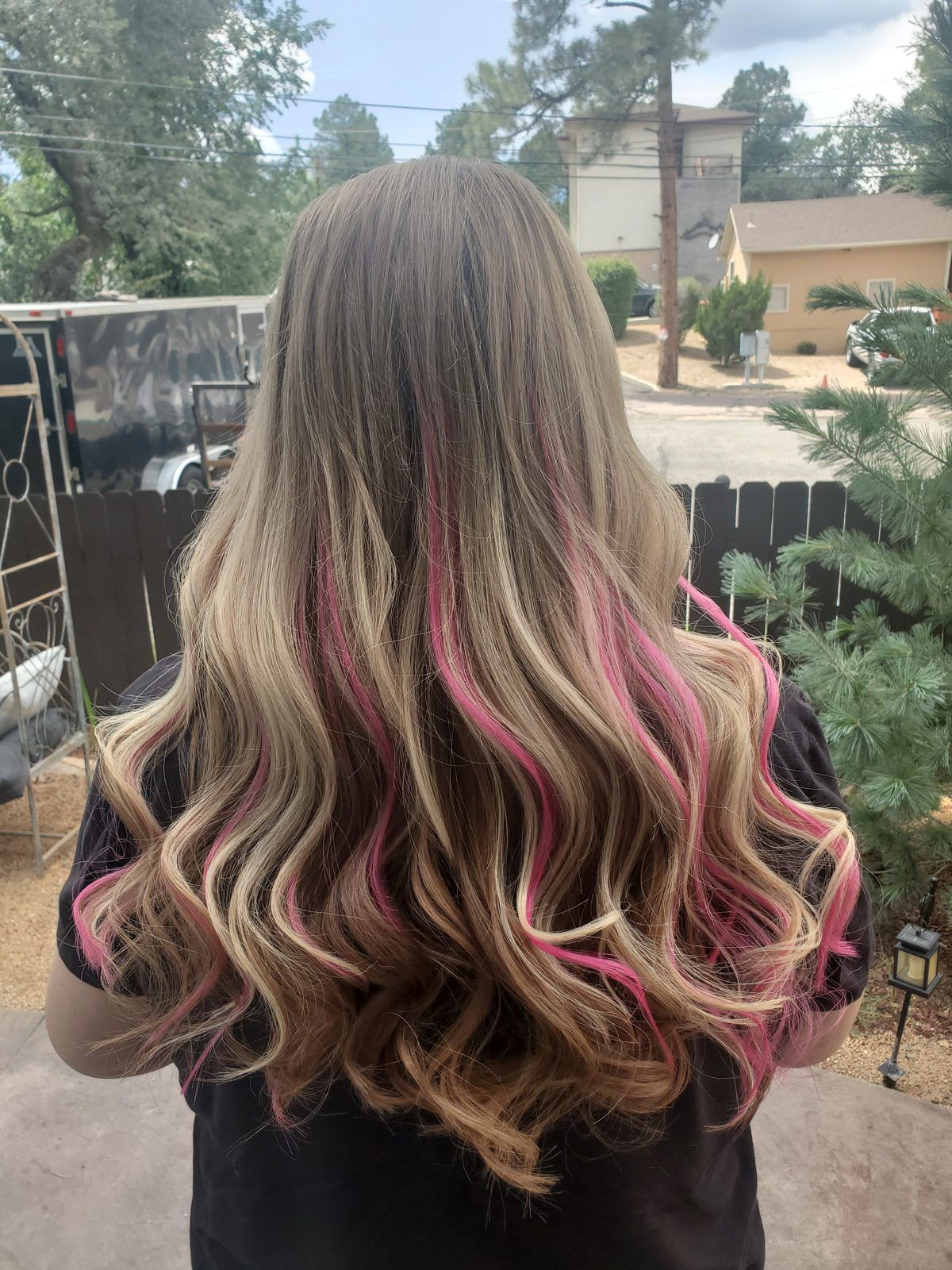 Long blonde waves with bright pink peekaboo streaks underneath, shown from behind outdoors.